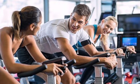 Young people talking and smiling while working out on bike at gym. Friends in a conversation while cycling on stationary bike in fitness centre. Group of happy people working out at spinning class.